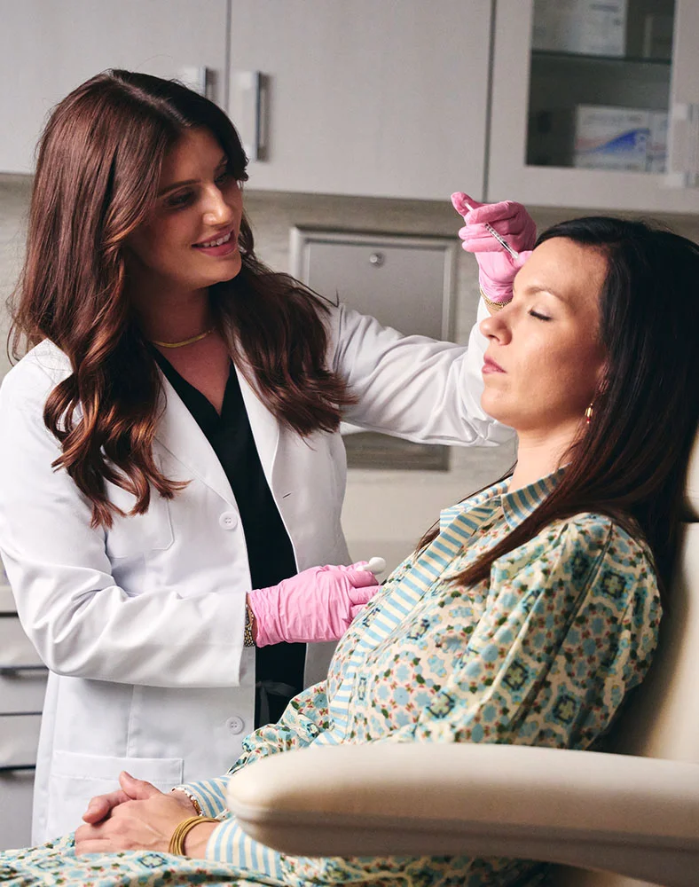 A female medical professional with long, dark hair, wearing a white lab coat and pink gloves, is administering an injection to a female patient who is seated in a chair with her eyes closed. The medical professional is smiling and looking at the patient, while the patient is relaxed and calm. The setting appears to be a medical office or spa. - Botoc and Wrinkle Relaxers in Macon, GA