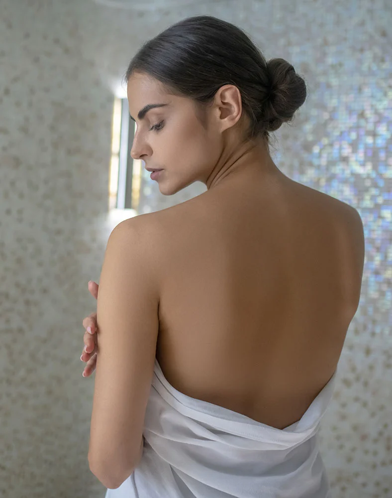 A young woman is shown from the back and side, wrapped in a white towel. Her dark hair is pulled back in a bun, revealing her bare shoulders and back. She is looking down and to the side, with a serene expression on her face. A mosaic-tiled wall is visible in the background. - Bra line Back Lift in Macon, GA