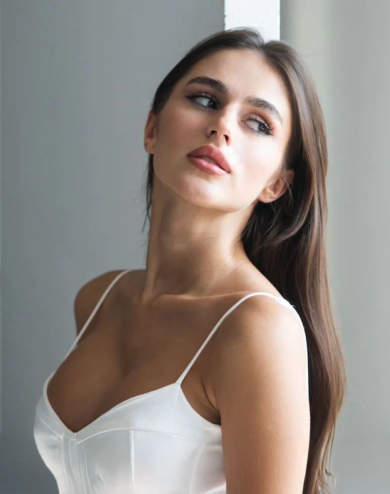 A beautiful young woman with long, dark hair is looking to the side. She is wearing a white satin camisole with thin straps. The image has a soft, diffused light, and she appears to be indoors with a gray wall as the backdrop. Her expression is thoughtful and serene. - Breast Reduction in Macon, GA