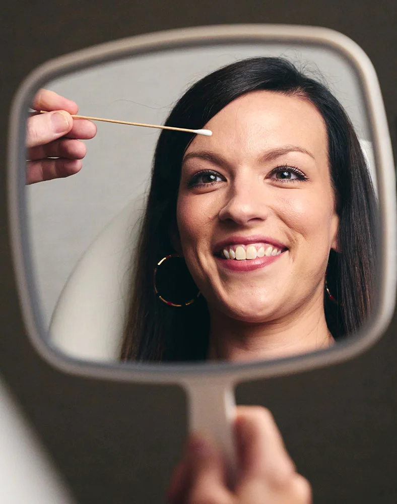 A mirror reflects a smiling woman with long, dark hair, wearing hoop earrings. A hand is reflected in the mirror, holding a cotton swab and touching the skin just above her eyebrow. The woman's face is in soft focus as she looks forward with a joyful expression. - Brow Lift in Macon, GA