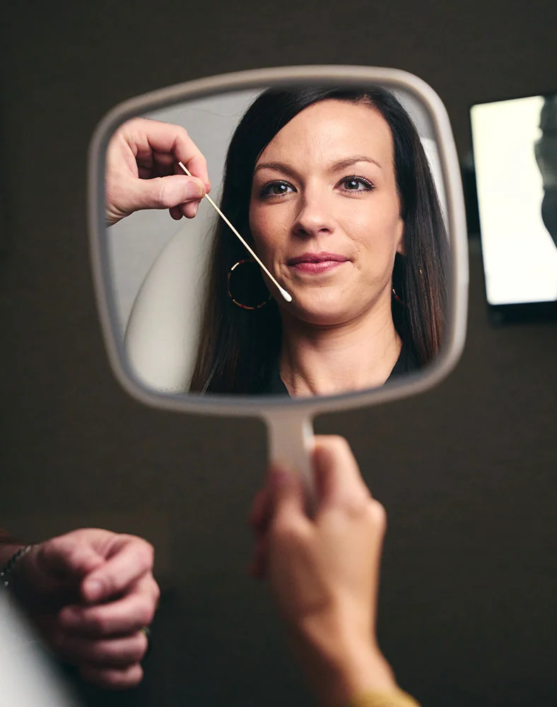 A mirror reflects the face of a woman with long, dark hair, wearing a black top and hoop earrings. Her face is in soft focus as she looks forward. A hand is reflected in the mirror, holding a long cotton swab and lightly touching the skin below her right eye. - Chin Implant in Macon, GA