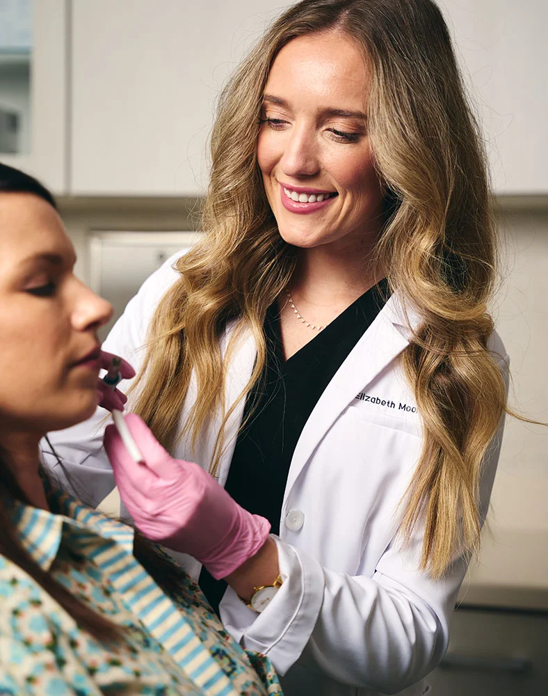 Close-up of a blonde female medical professional in a white lab coat and pink gloves performing a cosmetic lip procedure on a light-skinned patient wearing a patterned blouse. A thin white instrument is visible against a neutral-toned clinic background. - Dermal Fillers in Macon, GA