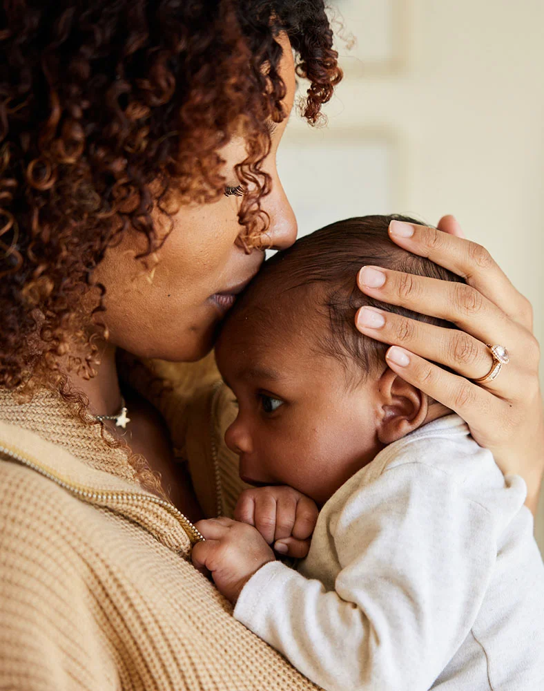 Close-up of a dark-skinned woman with curly dark brown hair cradling a baby. She wears a light beige textured sweater, a silver star-shaped pendant, and a small diamond-like ring. Her head rests gently against the baby’s forehead. The baby, dressed in a light gray or white garment, looks off to the side while the woman holds them close, emphasizing a tender, intimate moment. - Earwell in Macon, GA