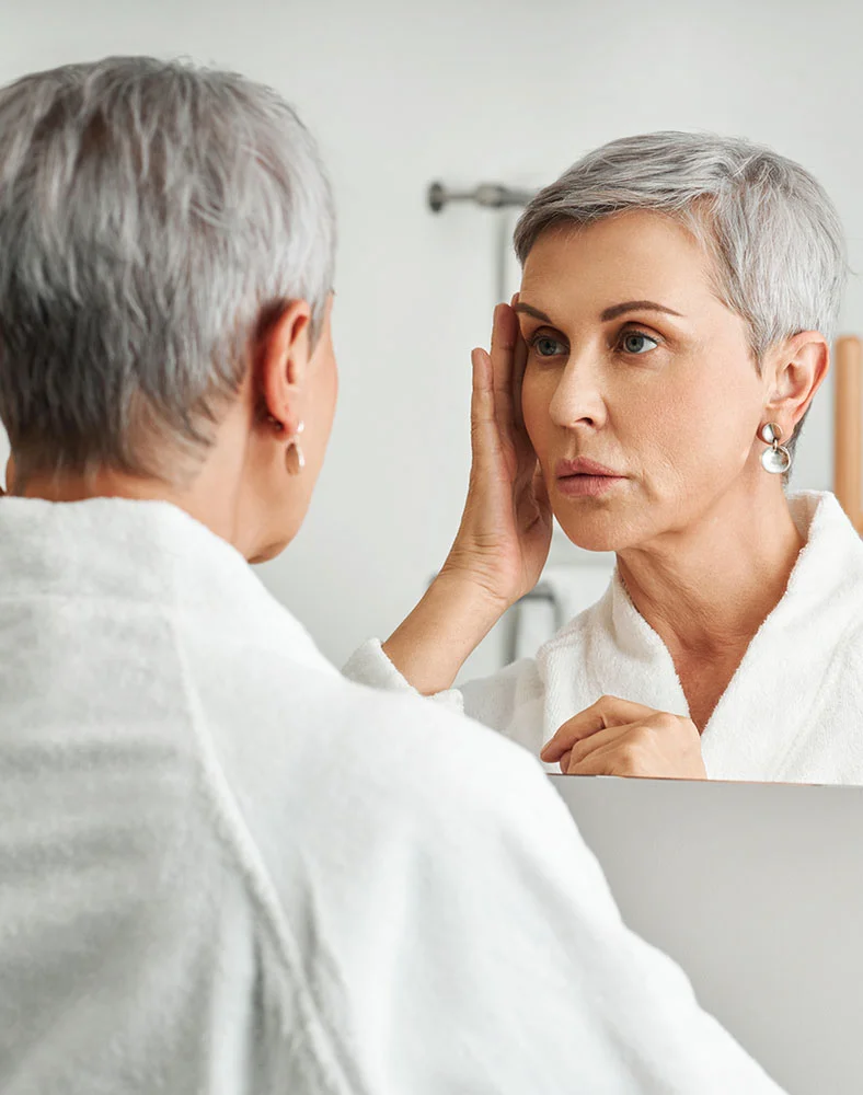A woman with short, gray hair is looking at her reflection in a mirror. She is wearing a white bathrobe. She is touching her face, and her expression appears somewhat concerned or pensive. The lighting in the bathroom is bright and neutral. The background is a light gray/white bathroom wall. The reflection in the mirror shows a similar-aged woman with a similar expression. - Eyelid Surgery in Macon, GA