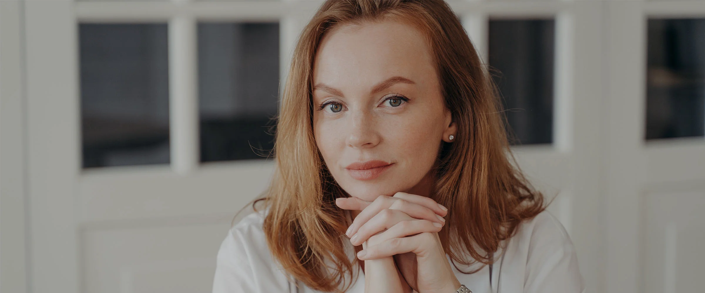 A light-skinned woman with shoulder-length reddish-brown hair is seated, looking directly at the camera with a thoughtful or serious expression. Her light-colored eyes are focused, and her hands are clasped in front of her chin. She wears a white long-sleeved button-up shirt over a dark top, with a silver-toned watch on her right wrist. The softly lit background is out of focus, featuring off-white walls or a light-colored room. - Face Lift in Macon, GA