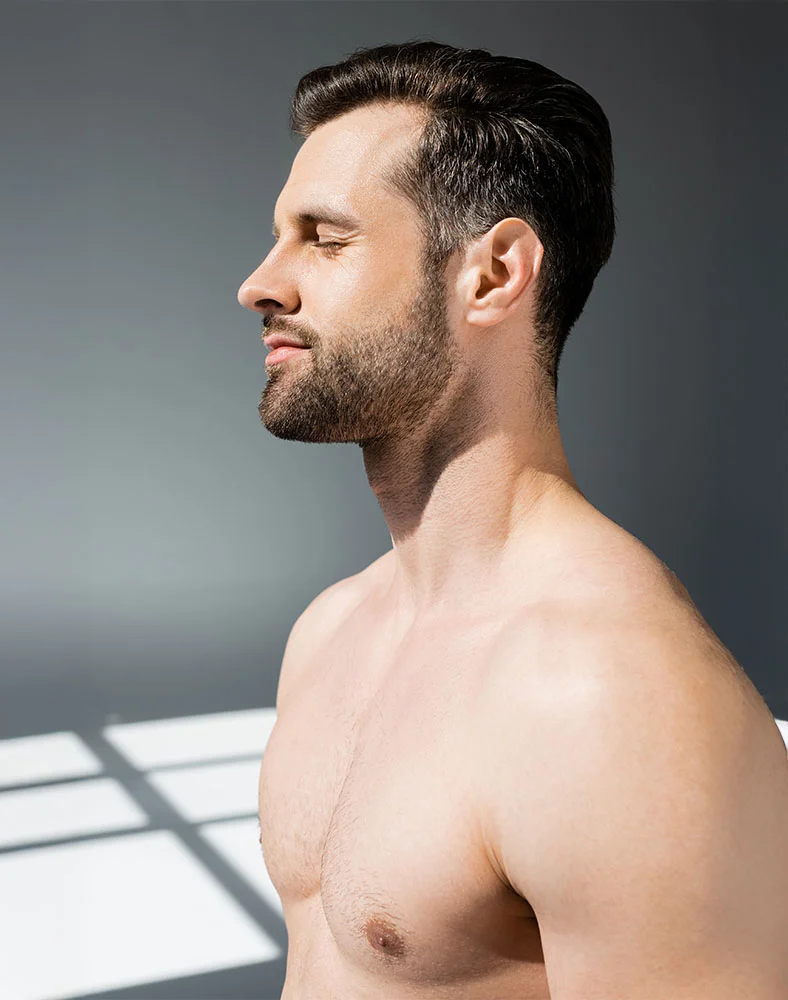 Profile view of a light-skinned, shirtless man with short, neatly styled dark hair and a short trimmed beard. His eyes are closed, and his expression is neutral and contemplative. His chest and shoulders show smooth, light skin with fine chest hair. The muted gray background features shadows cast by a light source, highlighting the contours of his face and body. - Gynecomastia in Macon, GA