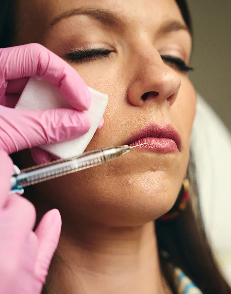Close-up of a woman receiving a cosmetic injection. A person wearing pink gloves holds a syringe and uses a cotton swab to clean the woman’s lower lip area. Her eyes are closed, appearing relaxed, while the focus stays on her face and the gloved hands preparing the injection against a softly blurred background. - Lip Fillers in Macon, GA