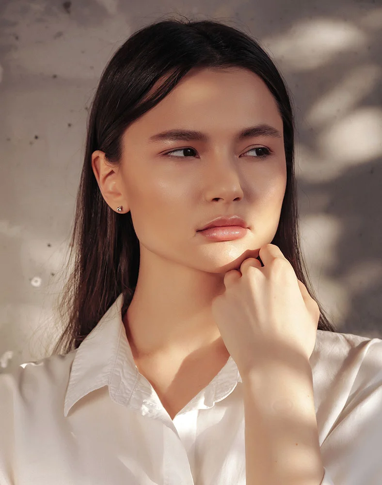 A woman with dark brown hair is seen from the mid-chest up against a textured light gray concrete wall. Her head is turned slightly to the right as she gazes into the distance. Dramatic lighting casts strong highlights on her face and hair, emphasizing her smooth, light-toned skin and subtle, natural makeup with rosy-pink lips. She appears bare-shouldered. - Microneedling in Macon, GA