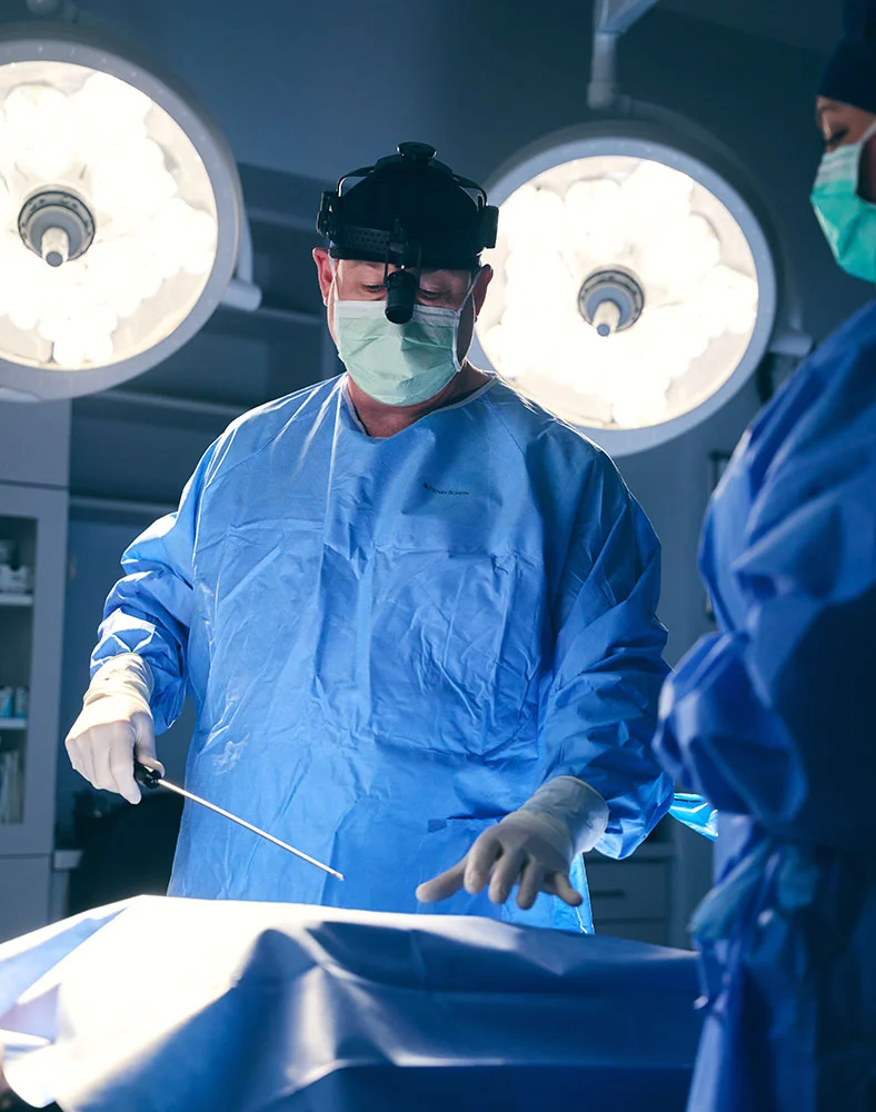 A close-up of a surgeon, Dr. Groves, in a blue gown, face mask, and headlamp, performing a procedure. He is holding a surgical instrument, with his gloved hands visible. Another person in blue scrubs is partially visible on the right. The background shows three large surgical lights and a sterile operating room environment. - Mommy Makeover in Macon, GA
