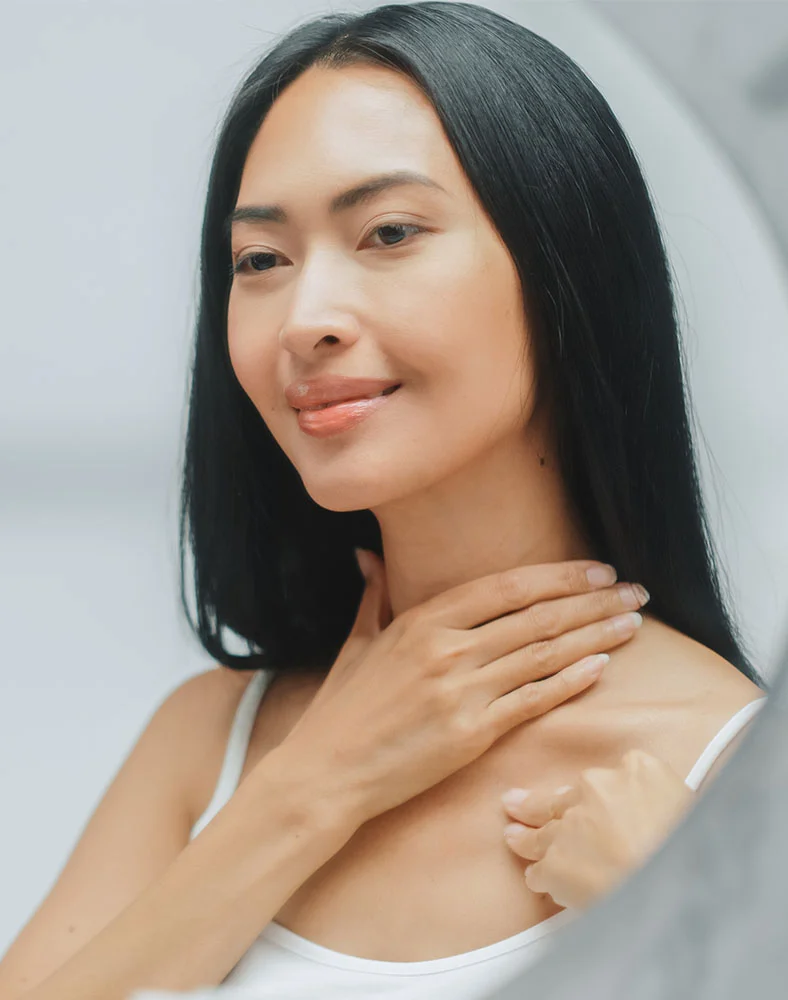 A young woman with long, straight black hair is shown in a mirror, looking at her reflection. She is smiling and touching the side of her neck with one hand. She is wearing a white tank top. The background is blurry and out of focus. - NeckLift in Macon, GA