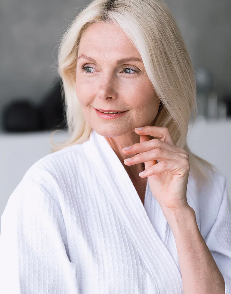A middle-aged woman with blonde hair and a white bathrobe is looking away from the camera and smiling. Her head is tilted slightly and she is resting her chin on her hand. The background is blurred, suggesting an indoor setting like a spa or hotel room. The lighting is soft and natural. - Vein Treatment and Therapy in Macon, GA